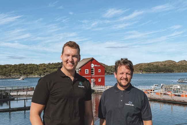 campusbla To menn som smiler foran sjø og blå himmel.