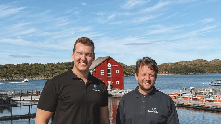 To menn som smiler foran sjø og blå himmel.