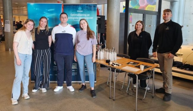 Seks menn og damer foran reklameplakat.