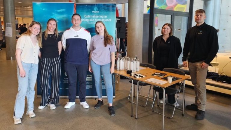 Seks menn og damer foran reklameplakat.